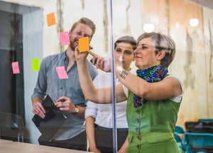 Three people review sticky notes on a glass wall in a conference room